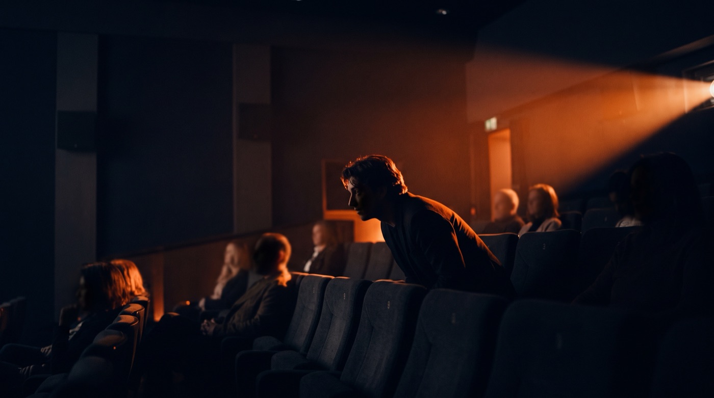 A small cinema audience in silhouette, one viewer leaning forward in warm projector light