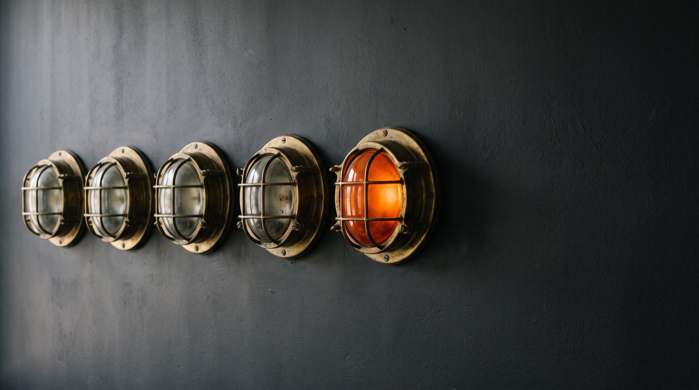 A row of five brass wall lamps on a charcoal wall, only the last one glowing orange